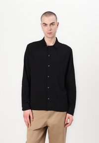Schwarze Hemdbluse mit langen Ärmeln, weicher Textur, klassischem Kragen und dunklen Knöpfen. Getragen über hellbraunen Hosen vor einem neutralen Hintergrund.