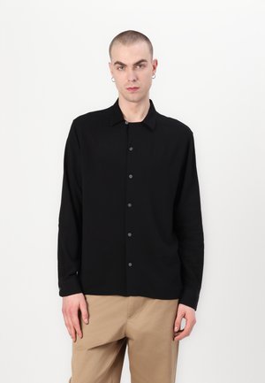 Black button-up shirt with long sleeves, soft texture, classic collar, and dark buttons. Worn over tan trousers in a neutral background.