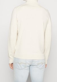 Person som bär en krämfärgad polo-tröja och ljusblå jeans, vänd bortom och visar detaljerna på baksidan av båda plaggen.