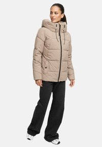 Beige pufferjack met een hoge kraag, ritssluiting aan de voorkant en elastische trekkoorden. Gecombineerd met zwarte wijde broek en witte sneakers.