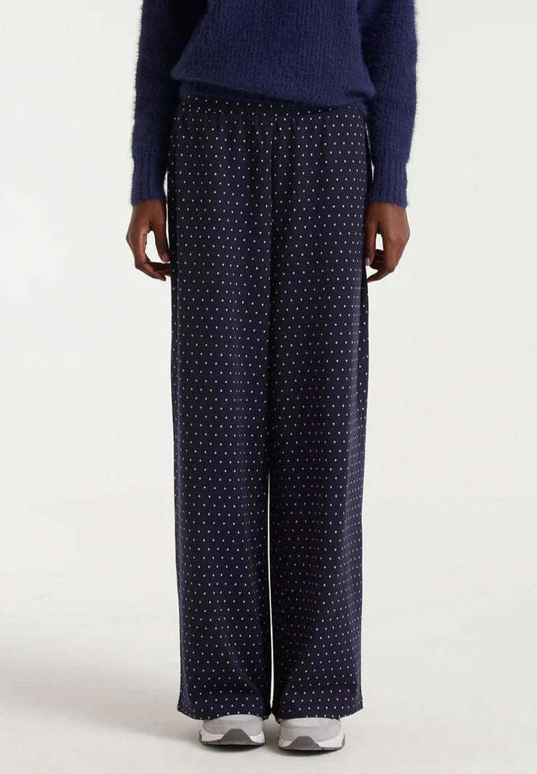 Persona vestida con pantalones anchos de color azul marino con pequeños lunares blancos, un suéter oscuro y esponjoso de color azul, y zapatillas blancas, de pie contra un fondo liso.