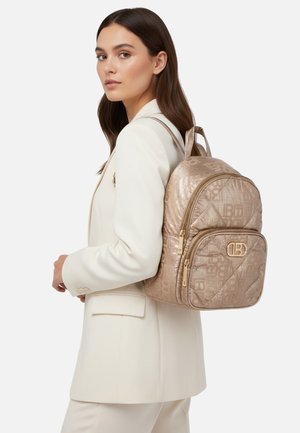 Zaino trapuntato beige con accessori dorati, caratterizzato da un logo prominente, doppie zip e due tasche. Texture liscia, design moderno.