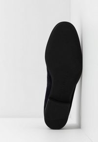 Semelle d'une chaussure de ville noire inclinée verticalement contre un coin de mur et de sol blancs, montrant des détails de bande de roulement lisse et texturée.