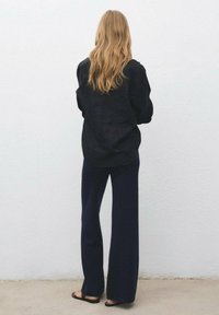 Una donna con lunghi capelli biondi mossi indossa un maglione nero largo, pantaloni navy a gamba larga e sandali neri, in piedi contro un muro bianco.