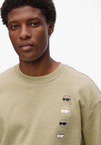 Sudadera de algodón verde oliva con cuello de canalé, que presenta cuatro gráficos de dibujos animados estilizados con gafas en negro, blanco y gris en el lado.