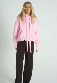 Růžová mikina s kapucí z měkké látky s přední kapsou, kapucí na šňůrku a vyšívaným nápisem; doplněná širokými tmavě hnědými kalhotami.