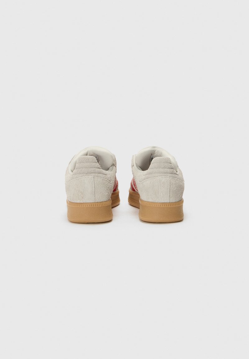 Des baskets avec une tige en daim blanc, des rayures en tissu rouge et une semelle en caoutchouc gum. Col et languette rembourrés avec des détails texturés à l'arrière.