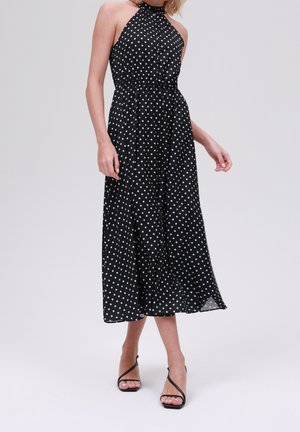 Czarna sukienka midi na szyję typu halter z białymi groszkami, elastyczną talią i zwiewną spódnicą, noszona z czarnymi sandałami na wysokim obcasie z paskami.