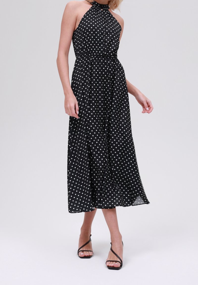 Robe midi noire à col licou avec pois blancs, taille élastique, jupe fluide, portée avec des sandales noires à talons hauts et à brides.