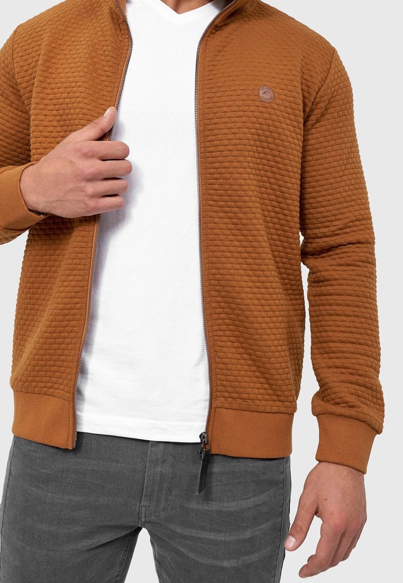 Homme portant une veste marron texturée zippée sur un t-shirt blanc et un jean gris, ajustant la fermeture éclair de la veste avec la main gauche.