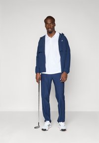 Námornícky golfový outfit s kapucňou, bielym polokošeľou a nohavicami. Biely golfový topánky so šedými akcentmi. Drží golfovú palicu.