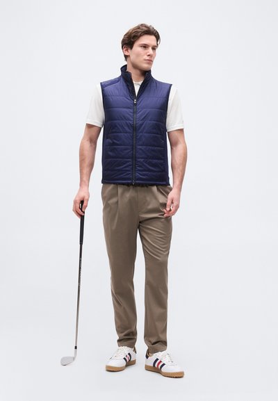 Άνδρας που στέκεται με ένα ρόπαλο γκολφ, φοράει γιλέκο σε σκούρο μπλε χρώμα με ραμμένες επενδύσεις, λευκό πουκάμισο με κοντά μανίκια, μπεζ παντελόνι και λευκά αθλητικά παπούτσια με ρίγες.