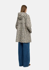 Beżowy płaszcz w czarne kropki, z kapturem, luźny krój i dużymi kieszeniami, zestawiony z szerokimi niebieskimi dżinsami.