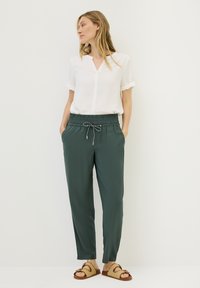 White short-sleeve blouse with a v-neckline, paired with dark green tapered trousers featuring an elastic waistband and drawstring.
