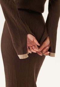 Robe marron côtelée avec de longues manches et de larges poignets beiges contrastés. Le tissu a une texture douce et une silhouette cintrée qui met en valeur la taille.