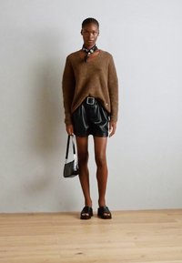 Bruine gebreide trui, zwarte leren shorts, zwarte sandalen en een kleine zwarte handtas. Een bandana om de nek voegt contrast toe. Eenvoudige, moderne stijl.