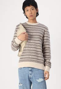 Suéter de punto en beige y marrón con patrones geométricos, puños y dobladillo acanalados. Combinado con jeans de corte desgastado en azul claro y un bolso beige.