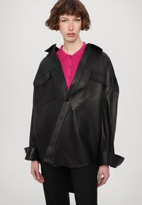 Jachetă din piele neagră, cu două buzunare frontale, guler deschis și croială oversized. Sub ea, se observă un top roz cu guler.