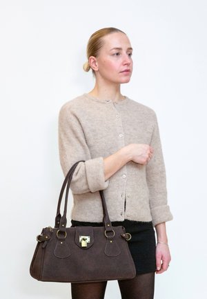 Frau mit blondem Haar, beige Cardigan und schwarzem Rock, die eine große dunkelbraune Handtasche mit goldenen Beschlägen hält und vor einem weißen Hintergrund steht.