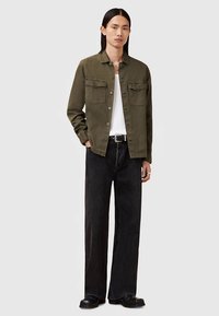 Oljčno zelena jakna z gumbi in dvema žepoma na prsih, pod njo bela majica, v kombinaciji s širokimi črnimi jeans hlačami in črnimi škornji.