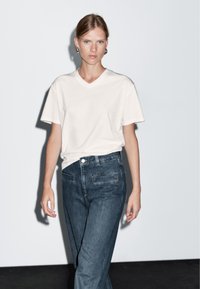 T-shirt bianco a scollo a V abbinato a jeans in denim scuro. Il t-shirt ha maniche corte e una vestibilità rilassata; i jeans presentano due tasche posteriori.