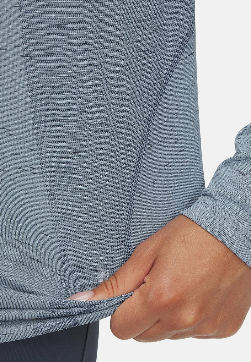 Lichtblauw sporttop gemaakt van gestructureerde stof, met een aansluitend ontwerp en lange mouwen met een subtiel patroon. Hand die de zoom vasthoudt.