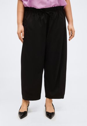 Άτομο που φορά χαλαρά μαύρα παντελόνια με ελαστική μέση και κορδόνι, συνδυασμένα με μαύρα παπούτσια με μυτερή μύτη και πουκάμισο μωβ που έχει μπει μέσα στο παντελόνι.