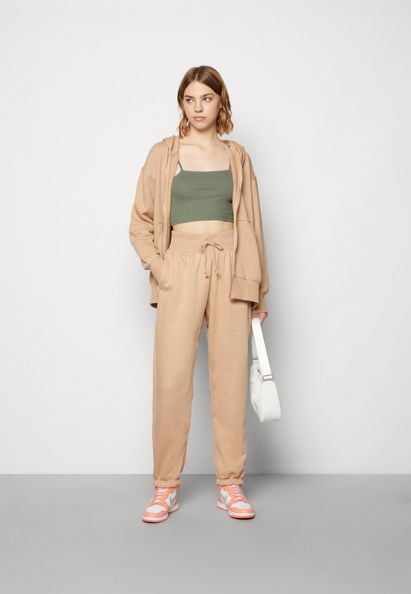 Felpa beige e pantaloni joggers con cordino, sovrapposti a un top verde corto. Sneakers rosa e grigie, e borsa bianca. Texture lisce.