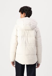 Veste rembourrée légère en crème avec capuche, présentant des sections matelassées horizontales et des accents élastiques au niveau des poignets. Texture lisse.