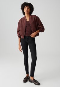 Femme aux cheveux bouclés portant une veste aviateur marron, un haut assorti, un jean skinny noir et des mocassins foncés, posant devant un fond uni.