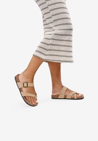 Sandales beige avec des brides réglables, semelle en liège et semelle en caoutchouc texturé. Les brides présentent un détail de boucle carrée.