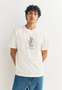T-shirt en coton blanc avec un motif de cactus et un texte en marron doux, col rond et manches courtes, porté avec un jean bleu.