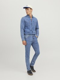 Camisa de denim azul claro con dos bolsillos en el pecho, combinada con jeans azules a juego. Zapatillas negras y una gorra de béisbol beige con acentos azules.