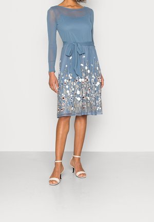 Kobieta ubrana w niebieską sukienkę do kolan z przezroczystymi długimi rękawami, kwiatowym haftem na spódnicy, przewiązana w pasie, nosząca białe sandały na wysokim obcasie.