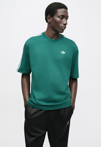 Zelené bavlnené tričko s okrúhlym výstrihom, krátkymi rukávmi a bielymi tromi pruhmi na ramenách. Na hrudi je malé biele logo.