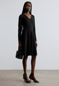 Must pikkade varrukatega kleit, millel on V-kaelus, volangseelik ja tekstuuriga kangas. Sobitatud mustade kontsakingade ja väikese musta käekotiga.