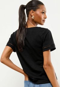 Melna īsām piedurknēm T-krekli ar brīvu piegriezumu, ar smalkām tekstūras akcentiem uz piedurknēm un apaļu kakla izgriezumu, apvienots ar gaišajiem džinsiem.