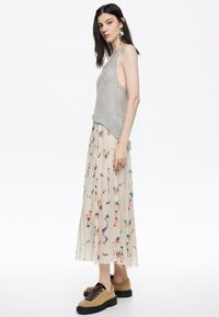 Top de tirantes de punto gris claro combinado con una falda plisada color crema con patrones florales multicolores. Zapatos marrones con cordones y suela gruesa.
