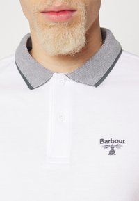 Gros plan d'une personne avec une barbe blond clair portant un polo blanc avec un col gris et le logo Barbour sur la poitrine.