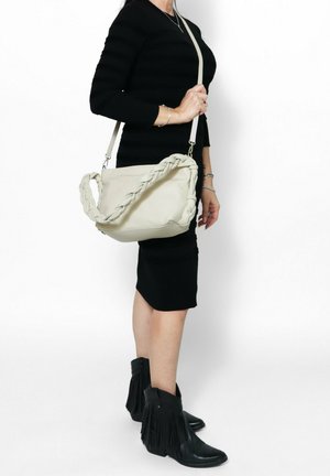 Borsa a tracolla in pelle beige con manico intrecciato, tracolla regolabile e superficie liscia, indossata da una persona che porta un abito nero.