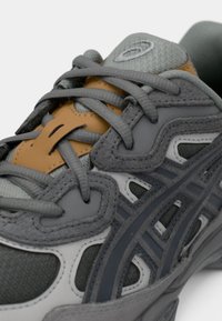 Γκρι και καφετί αθλητικό παπούτσι με υφασμάτινη υφή, επικολλημένα συνθετικά πάνελ και σταυρωτά γκρι κορδόνια πάνω από τη γλώσσα.