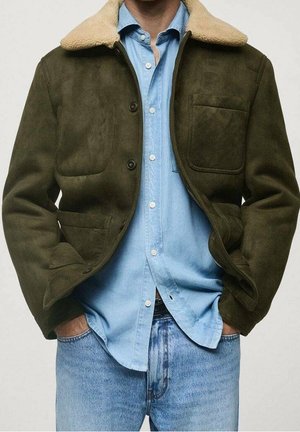 Mężczyzna ubrany w oliwkowo-zieloną kurtkę z futrzanym kołnierzem na jasnoniebieską koszulę z denimu i niebieskie jeansy, luźno trzymający ręce w kieszeniach, na jednolitym tle.