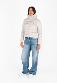 Lichtbeige pufferjack met een capuchon, met horizontale stiksels, een ritssluiting aan de voorkant en verstelbare trekkoorden. Gepaard met wijde blauwe jeans.
