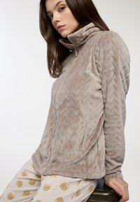 Giacca in pile beige soffice con zip e colletto alto, caratterizzata da un motivo a chevron testurizzato. Il modello indossa pantaloni da pigiama stampati con motivi di orsi.