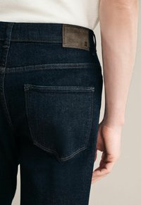 Tumed denimpüksid, millel on tagatasku, heledad õmblused ja tekstureeritud pruun nahkpakk vöö kohal.