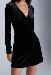 Vestido de terciopelo negro con manga larga, escote en V y detalle de cintura fruncida. Textura suave y dobladillo a la altura de la rodilla.