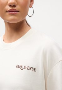 Plano cercano de una persona que lleva una camisa blanca con el texto "PARK AVENUE" y un pendiente de aro plateado, sobre un fondo blanco.