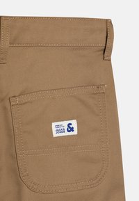 Beige puuvillased püksid, millel on taga tasku sinise ja valge sildiga. Kanga tekstuur on sile ja tugevdused on õmblustes.