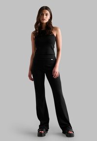 Schwarzes ärmelloses Top kombiniert mit schwarzen weit geschnittenen Hosen. Beide aus glattem Stoff mit minimalistischem Design und dezentem Logo-Detail. Schwarze Plateausandalen.