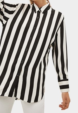 Femme portant une chemise à manches longues boutonnée avec de larges rayures verticales noires et blanches, associée à un pantalon blanc, bras et torse visibles.